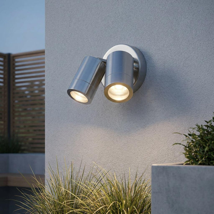 Illuminated twin stainless steel outdoor spotlight mounted on a textured gray wall above garden grasses.