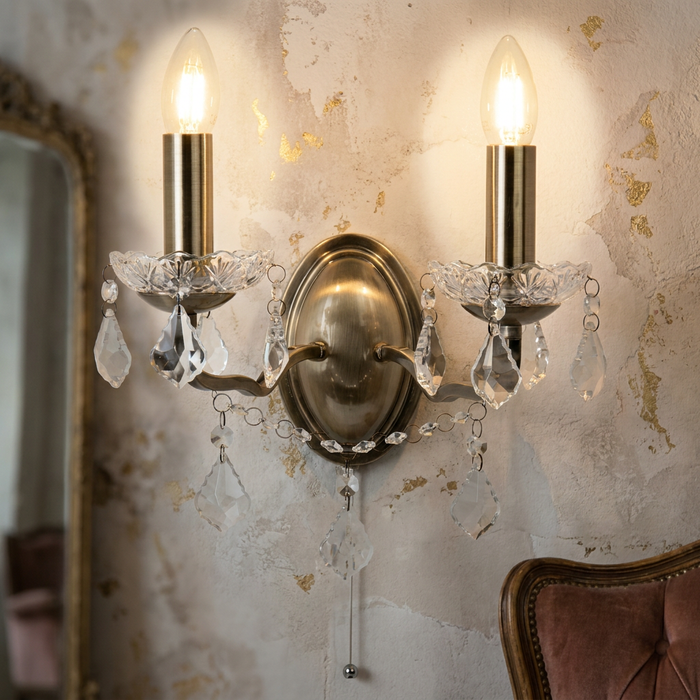 Two-light antique brass wall sconce with crystal drops and cord pull on a distressed textured wall.