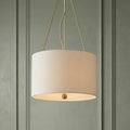 Cream linen drum pendant light with brass accents suspended in front of a sage green paneled wall.