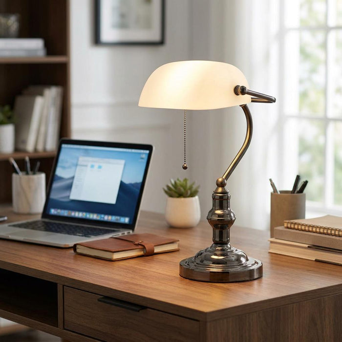 A bronze banker's lamp with a pearl glass shade lit on a wooden desk next to a laptop in a home office.
