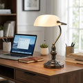 A bronze banker's lamp with a pearl glass shade lit on a wooden desk next to a laptop in a home office.