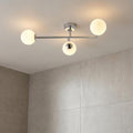Modern chrome three-light semi-flush bathroom fixture with glowing opal globe shades against light grey tiled walls.
