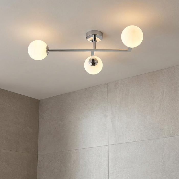 Modern chrome three-light semi-flush bathroom fixture with glowing opal globe shades against light grey tiled walls.