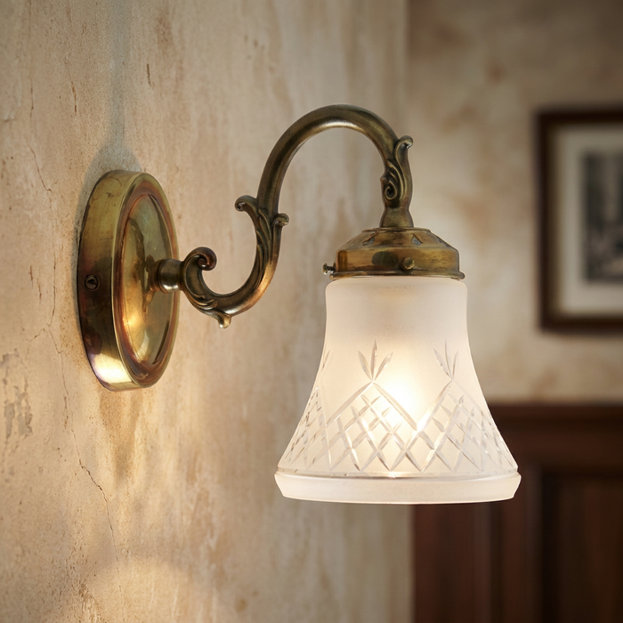 Pinestar Art Deco wall light with a brass swan-neck arm and frosted cut-glass shade mounted on a textured wall.