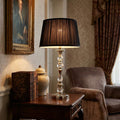 Polished nickel table lamp with glass accents and a black pleated shade on a dark wood side table with antique books.