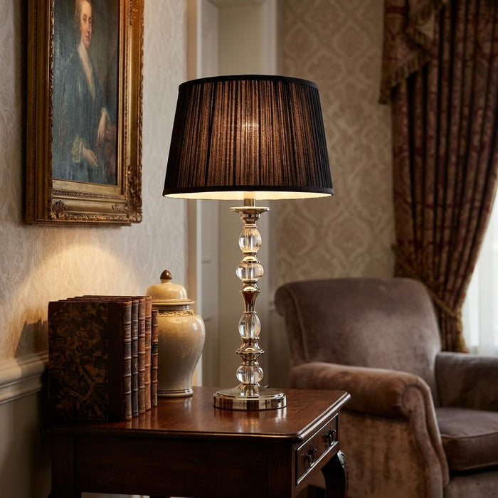 Polished nickel table lamp with glass accents and a black pleated shade on a dark wood side table with antique books.