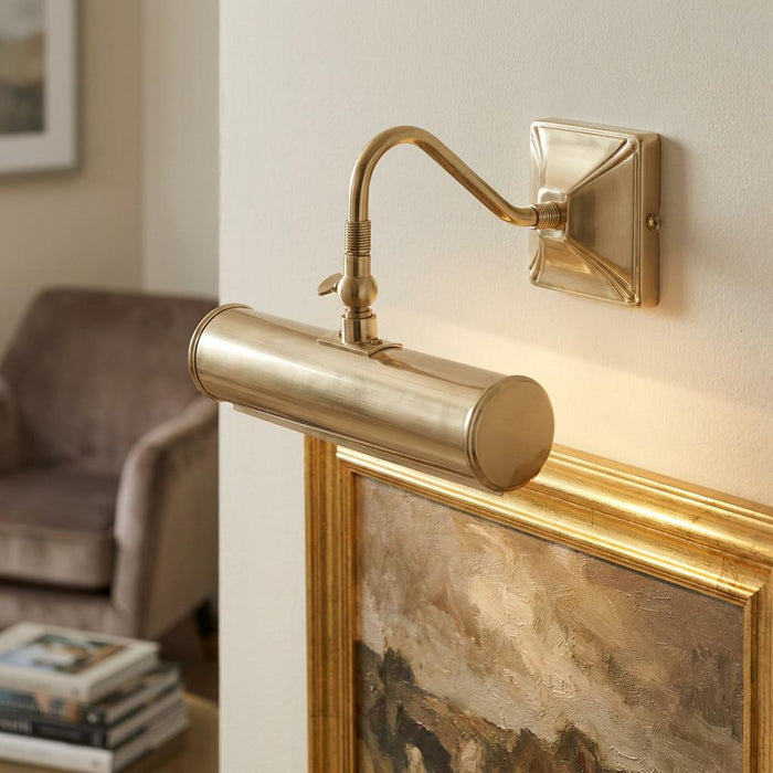 Polished brass picture light with an adjustable arm mounted on a wall above a gold-framed painting.