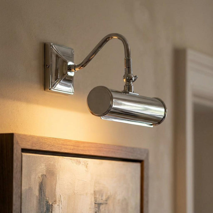 Polished chrome picture light with a curved arm illuminating a framed painting on a wall.