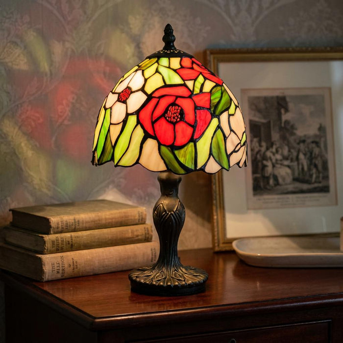 Lit Poppy Tiffany bedside lamp with a floral stained glass shade on a wooden table next to vintage books.