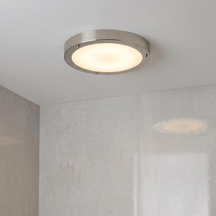 A circular brushed nickel and frosted glass flush-mount ceiling light illuminated on a white bathroom ceiling.