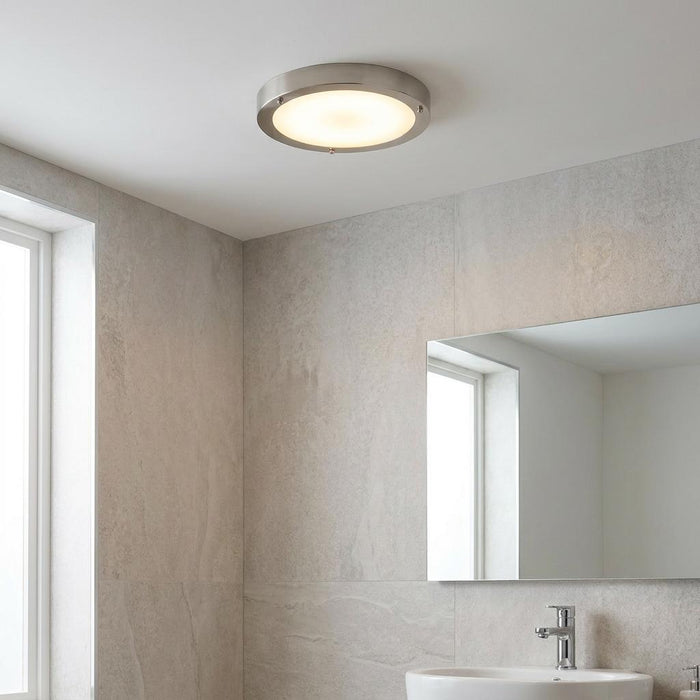 A circular brushed nickel flush bathroom ceiling light with frosted glass glows in a modern grey tiled bathroom.