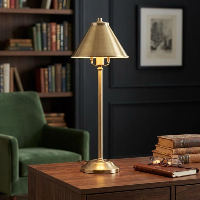 Brass stick table lamp with a cone shade on a wooden desk in a dimly lit library setting.