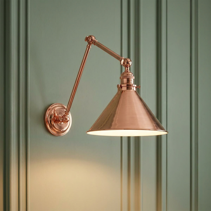 Polished copper adjustable swing arm wall light with a conical shade mounted on a green paneled wall.