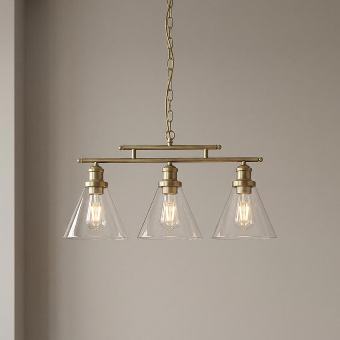 Antique brass 3-light ceiling pendant with clear conical glass shades and illuminated Edison bulbs on a plain wall.