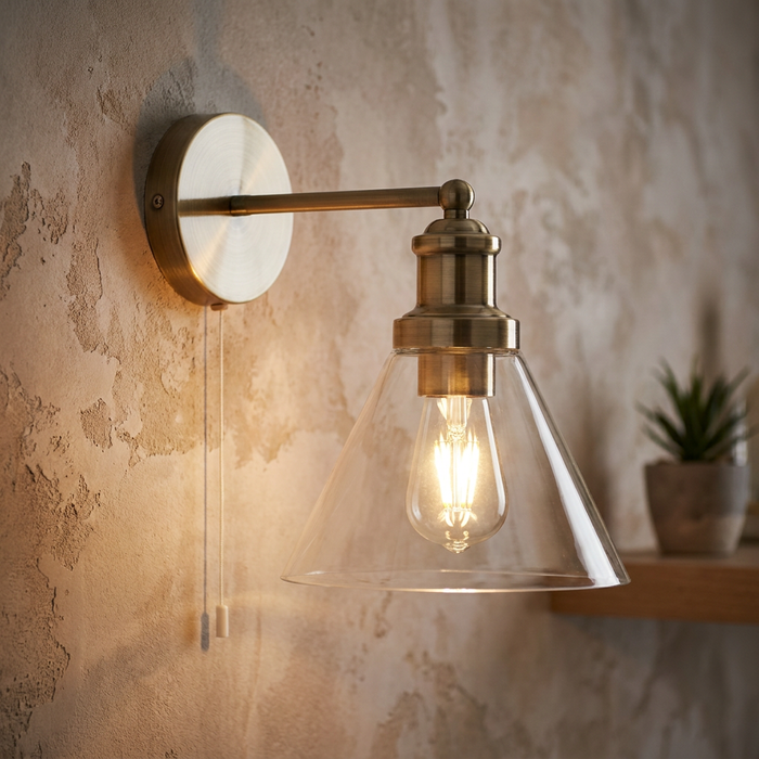 Pyramid brass wall light with a clear glass cone shade and a cord pull switch on a textured wall.