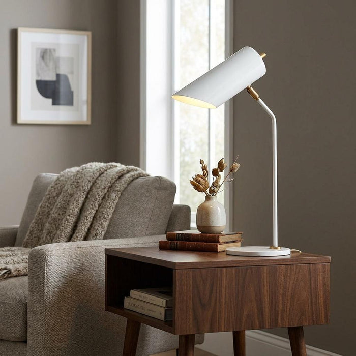 Modern white and aged brass table lamp illuminates a wooden side table next to a grey armchair with a cozy throw.