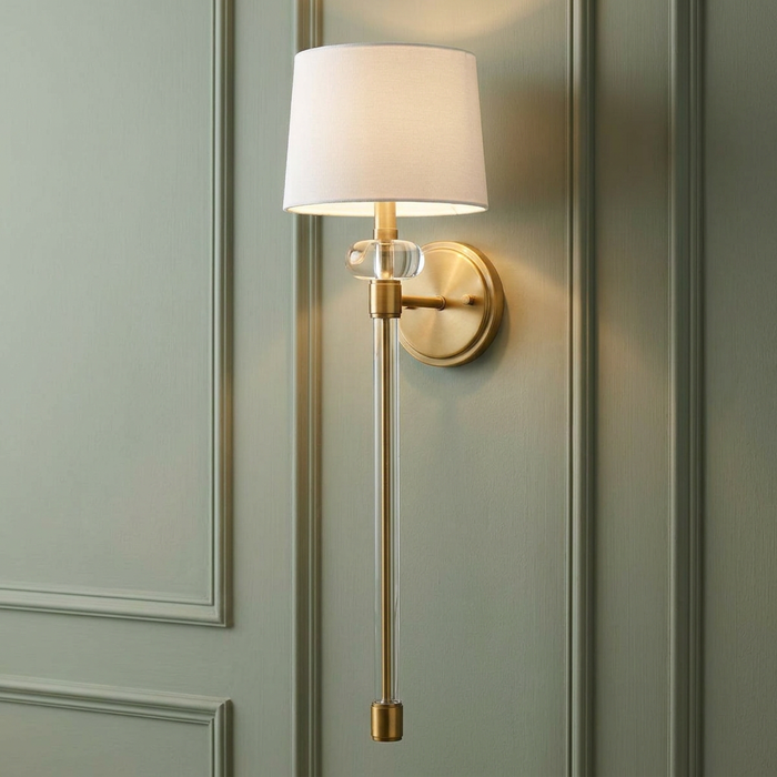 A brass wall sconce with a white fabric shade and long glass column mounted on a green paneled wall.