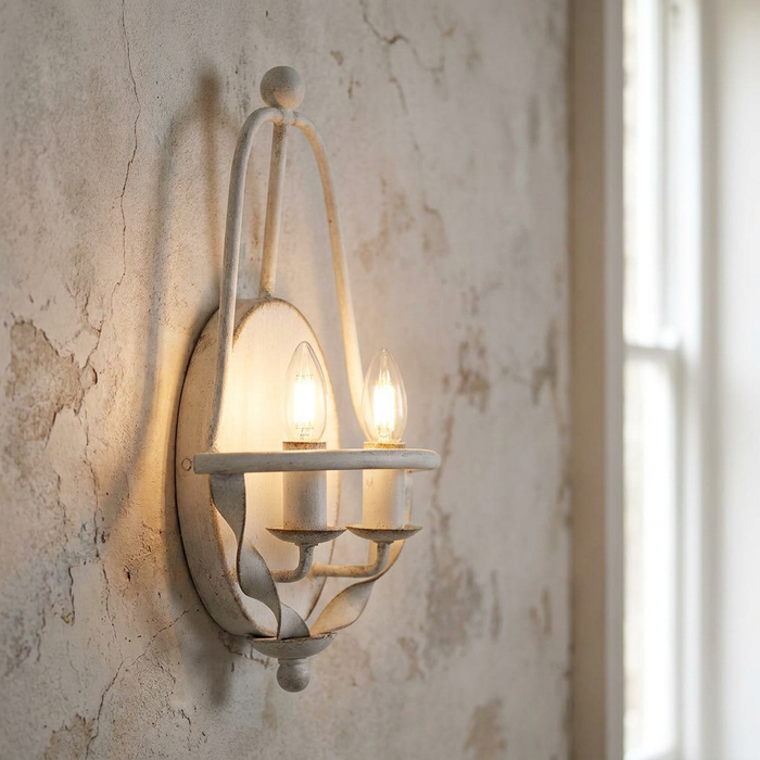 Distressed white two-light wall sconce with candle bulbs mounted on a rustic, weathered plaster wall.