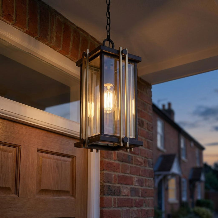 Quoizel Goldenrod bronze and gold outdoor pendant lantern hanging from a porch ceiling against a brick wall at dusk.
