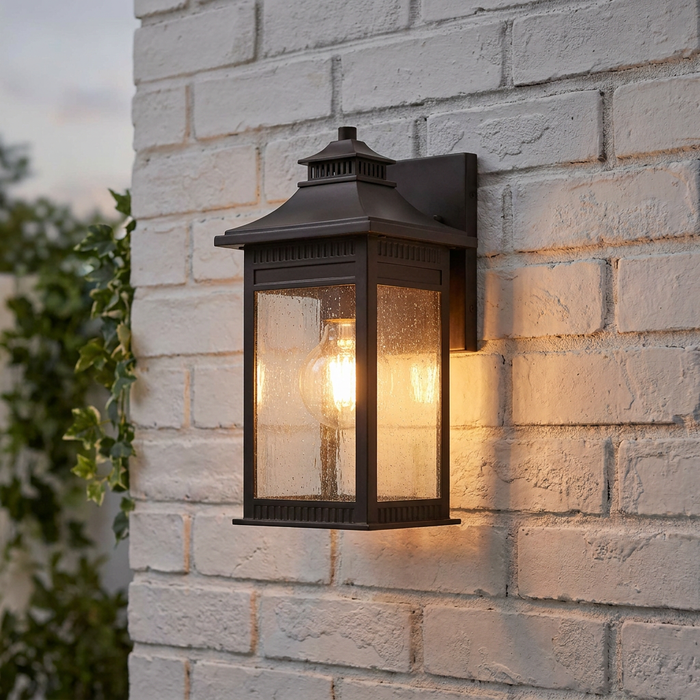 Bronze outdoor wall lantern with seeded glass panels glowing on a white brick wall.
