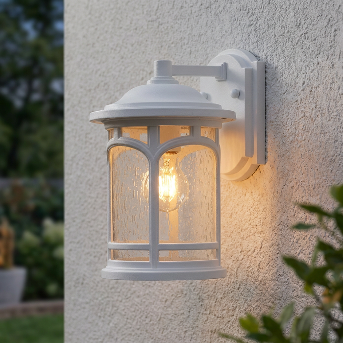 White traditional outdoor wall light with seeded glass and a warm glowing bulb on a textured stucco wall.