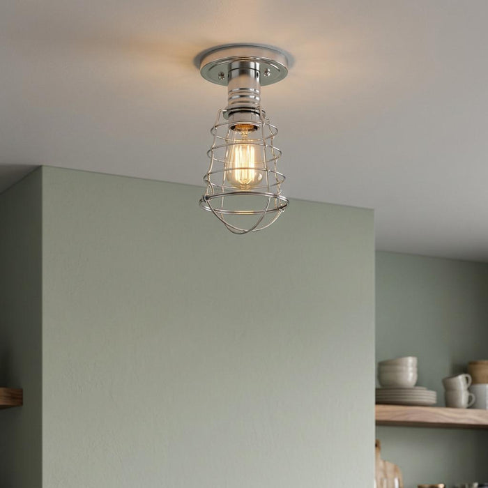 Industrial chrome wire cage semi-flush mount light with a glowing bulb against a light green wall.