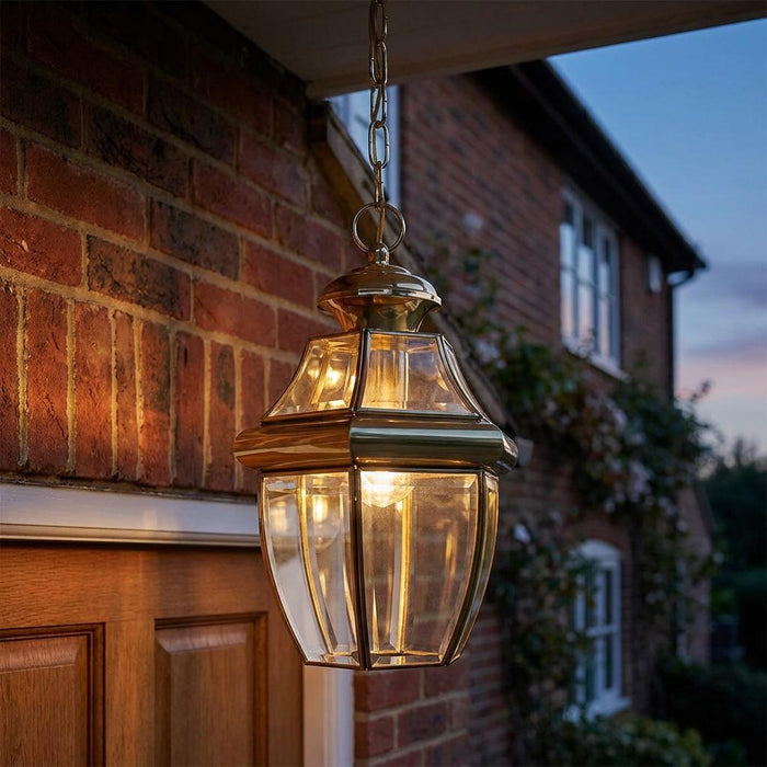 Lit polished brass chain lantern with clear glass panels hanging outside a brick house at twilight.