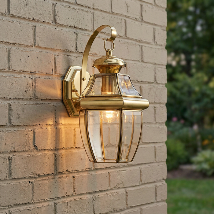 Traditional polished brass outdoor lantern with clear beveled glass panels glowing on a light brick wall.