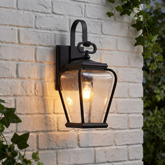 Black lantern wall light with seeded glass panels glowing on a white brick wall next to green ivy.