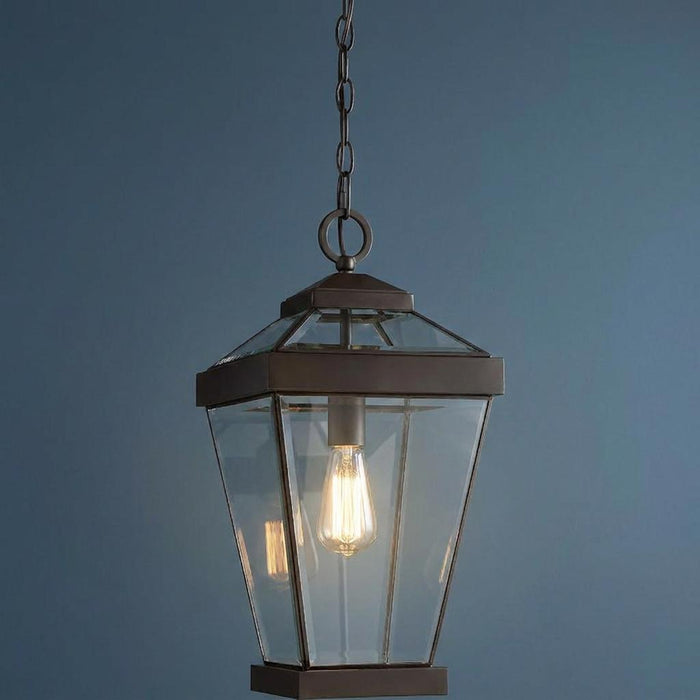 Bronze outdoor pendant lantern with clear glass panels and a lit Edison bulb against a blue wall.