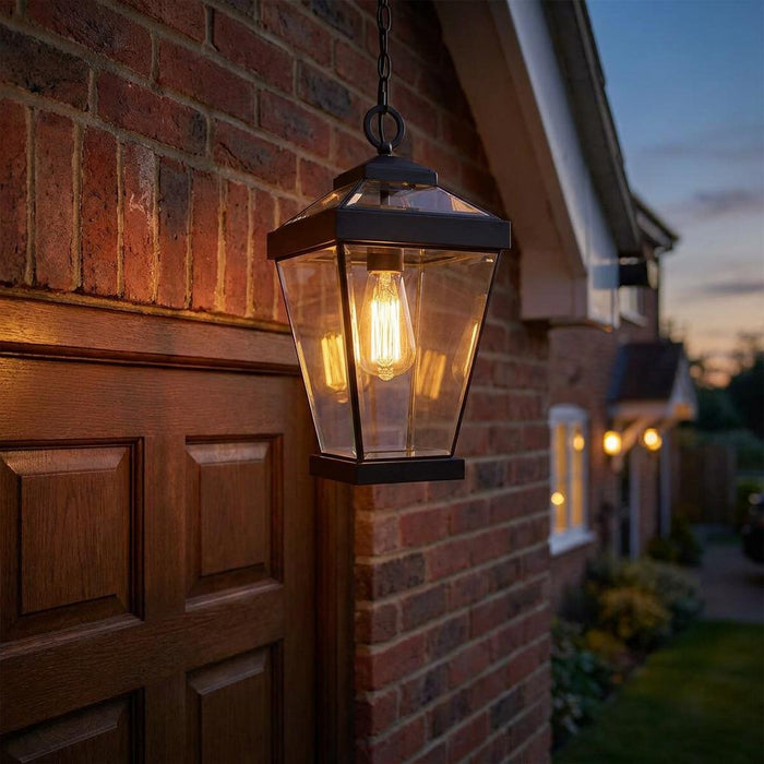 Glowing bronze outdoor chain lantern with clear glass panels hanging beside a brick wall and wooden door at dusk.