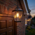 Glowing bronze outdoor chain lantern with clear glass panels hanging beside a brick wall and wooden door at dusk.