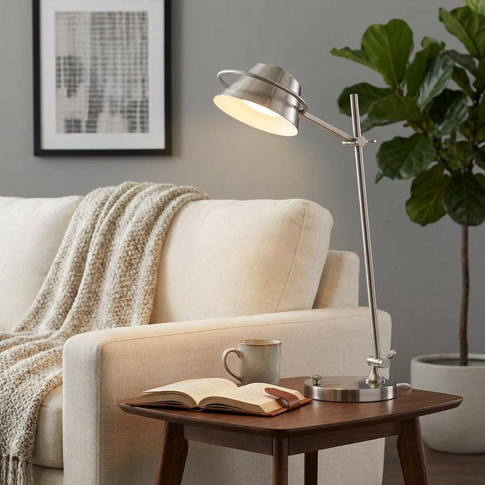 Brushed nickel LED table lamp illuminating an open book and mug on a wooden side table next to a beige sofa.