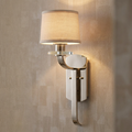 Polished silver wall sconce with a cream fabric shade and crystal accent, emitting a warm glow on a textured wall.