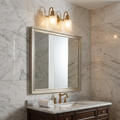 Brushed brass two-light wall sconce with frosted glass shades mounted above a silver mirror in a marble bathroom.