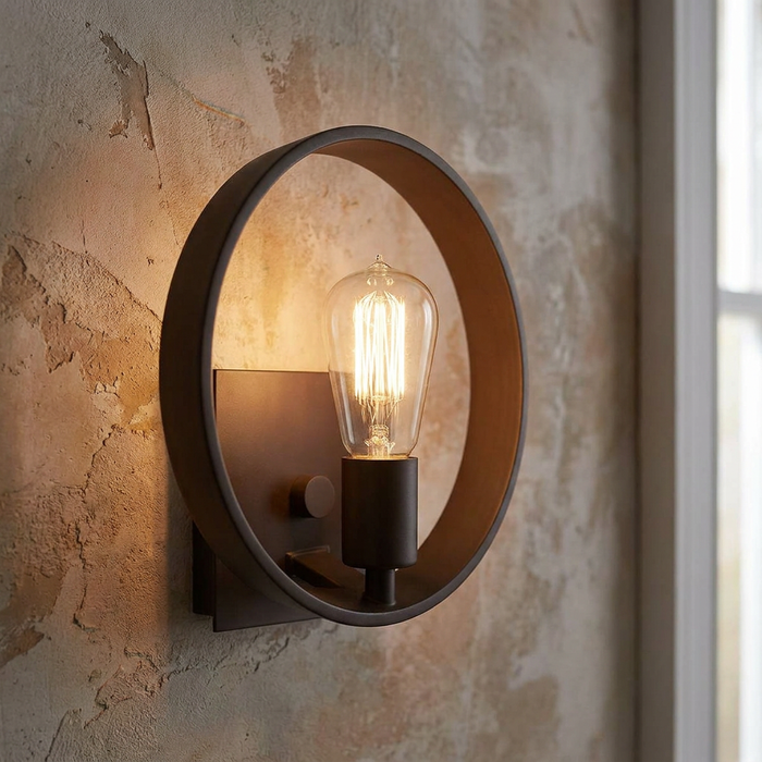 Industrial circular bronze wall sconce with a warm glowing Edison bulb against a textured plaster wall.