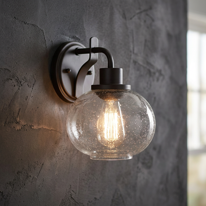 Wall-mounted light with a bronze finish, seeded glass globe shade, and a warm Edison bulb on a textured wall.