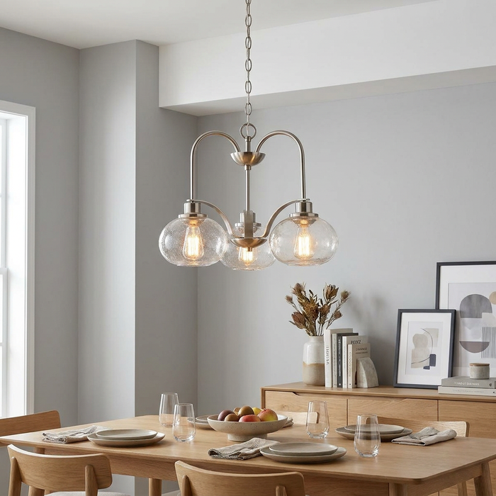 A 3-light brushed nickel chandelier with seeded glass globes hanging over a wooden dining table in a modern room.