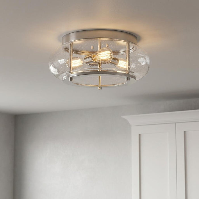 Brushed nickel flush mount ceiling light with a seeded glass shade and three glowing Edison bulbs in a neutral room.