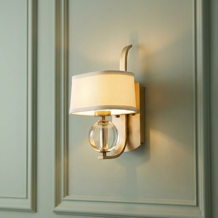 Modern brass wall light with a cream fabric shade and crystal sphere accent on a sage green paneled wall.