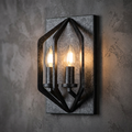 Industrial black metal wall sconce with a geometric cage and two lit candle bulbs on a textured grey wall.