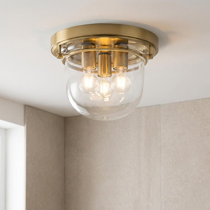 A three-light brass ceiling flush mount with a clear glass dome shade, illuminated in a neutral bathroom.