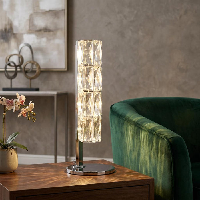 Remy LED chrome and hexagonal crystal table lamp illuminated on a wooden table beside a green velvet armchair.
