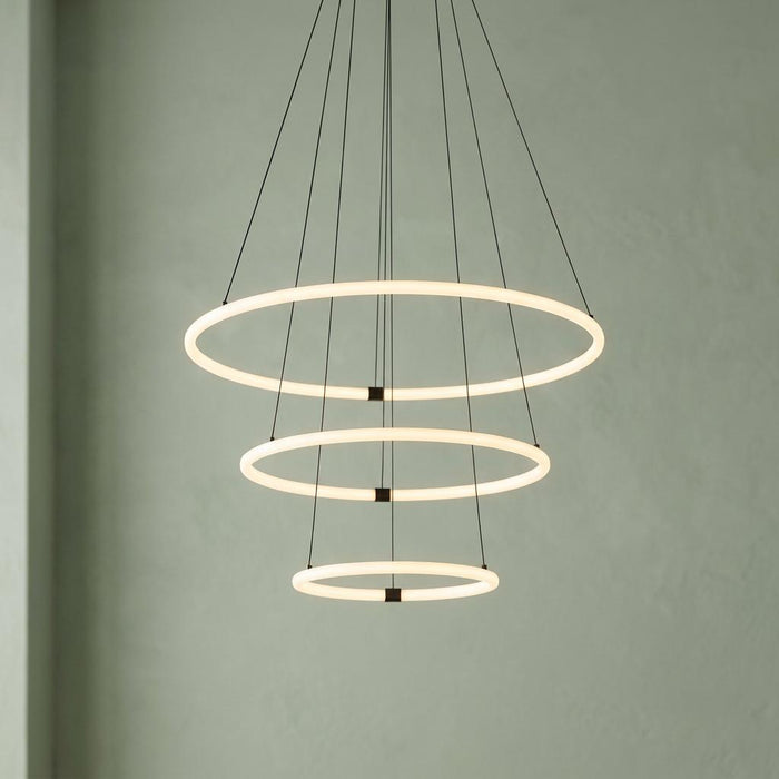 A three-tiered LED ring pendant light with glowing opal tubes suspended by black wires against a light green wall.
