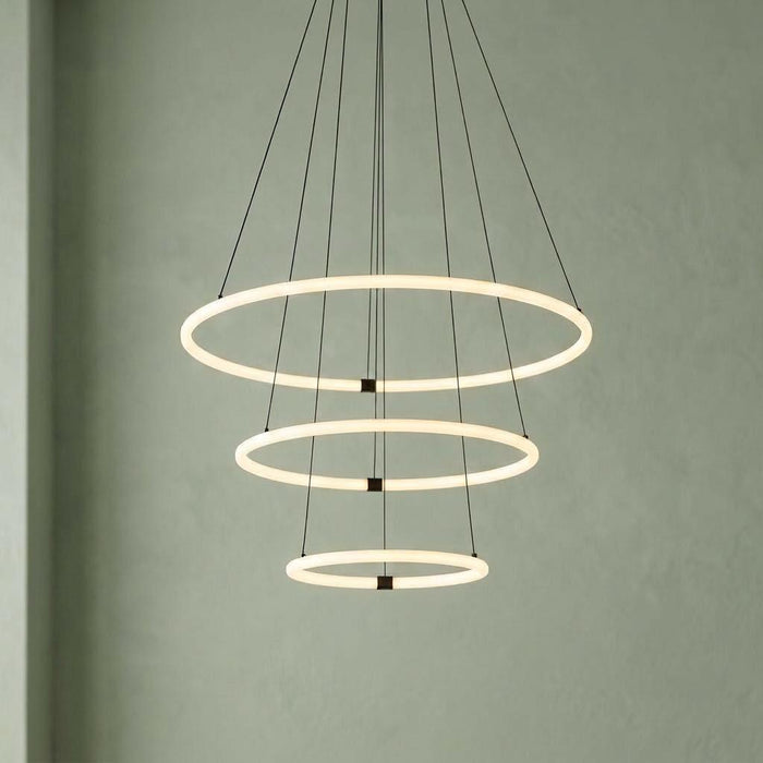 Three-tier LED ring pendant light with opal tubes suspended by black cables against a soft green wall.
