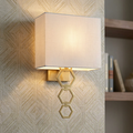 Aged brass wall light with a white fabric shade and three interlocking hexagonal rings on textured wallpaper.