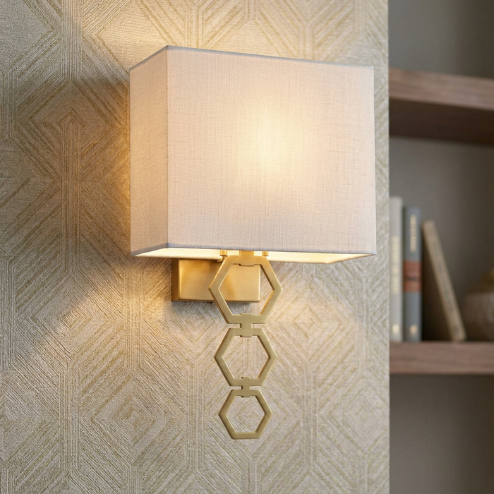 Aged brass wall light with a white fabric shade and three interlocking hexagonal rings on textured wallpaper.
