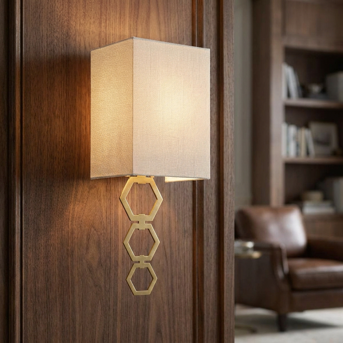 Aged brass wall sconce with a rectangular fabric shade and decorative hexagonal links on a dark wood wall.