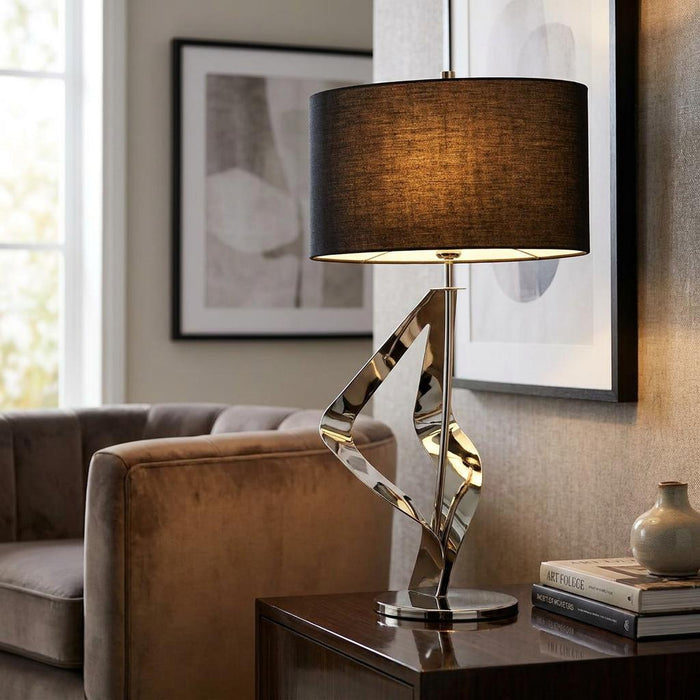 Modern table lamp with a black drum shade and polished chrome ribbon base on a side table.