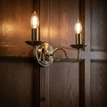 Brass two-light scroll arm wall sconce with lit candle bulbs and a pull cord, mounted on dark wood paneling.